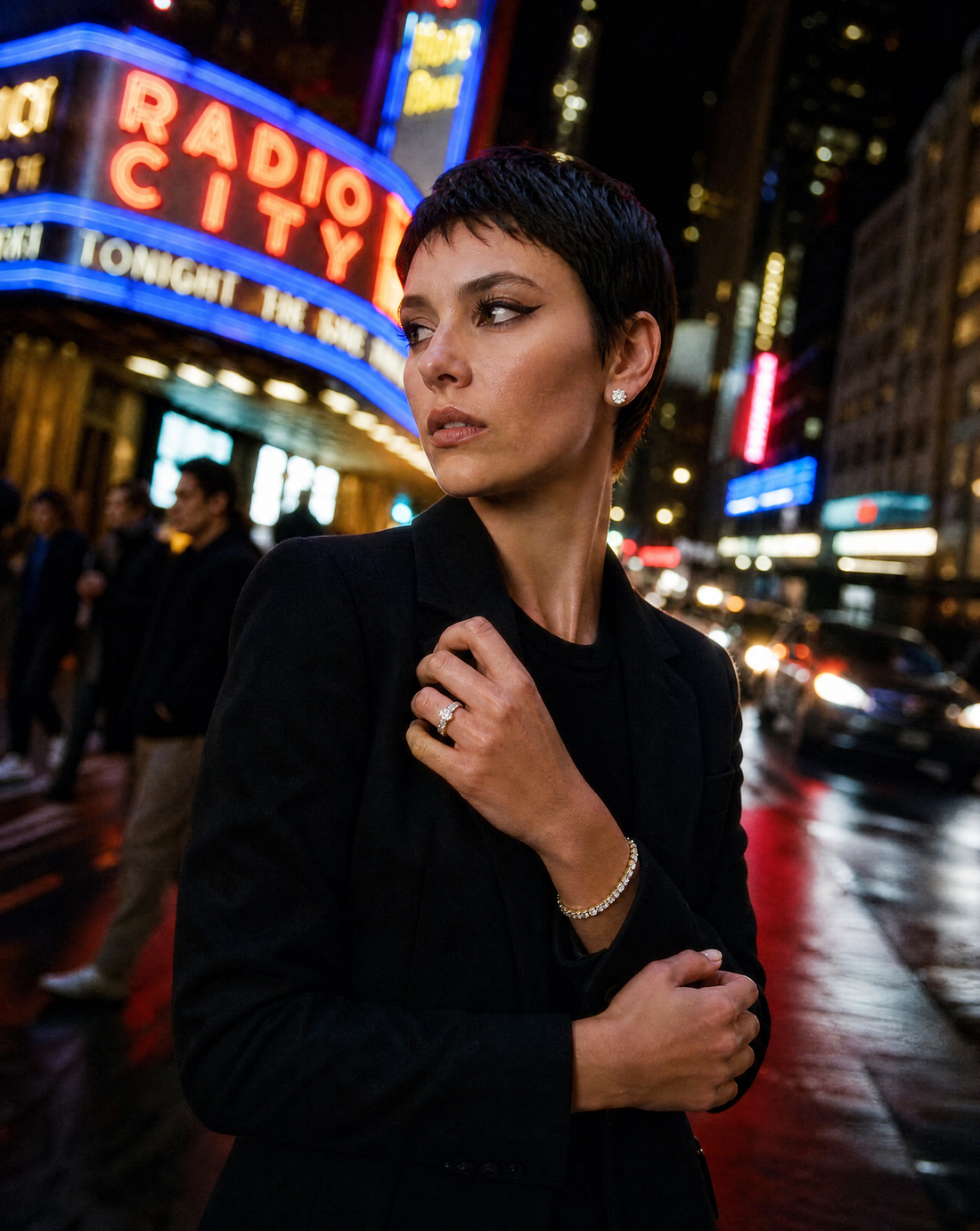Woman in a black blazer wearing a gold tennis bracelet, diamond studs and a ring on a city street at night