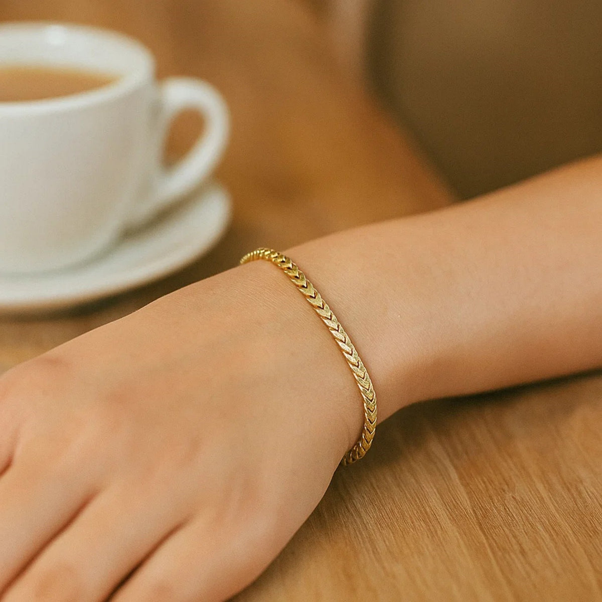 Gold bracelet worn on a wrist beside a coffee cup