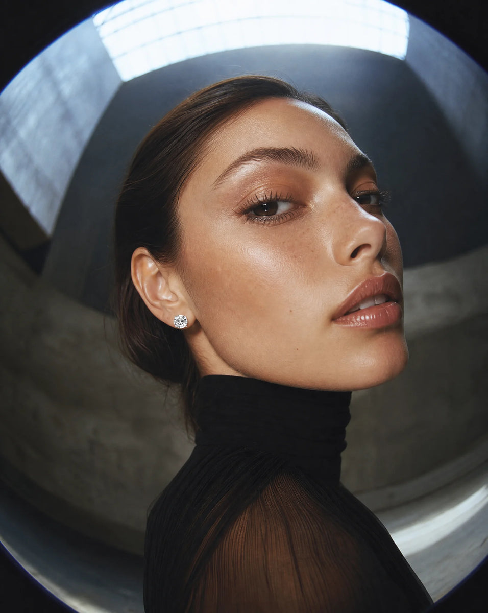 Close-up of a person wearing 2 carat IGI certified lab grown diamond stud earrings in 14k solid gold  a black turtleneck with a blurred background