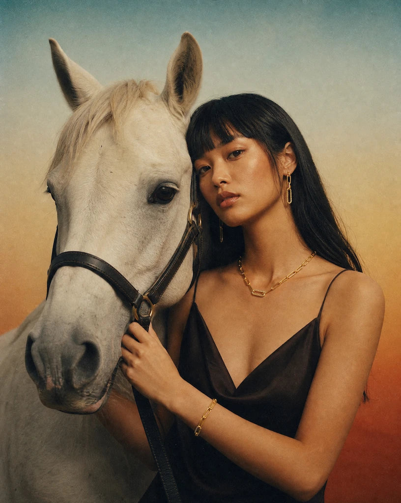 A model wearing solid 14K gold jewelry set from Deeve Canada in a black dress standing next to a white horse with a warm, gradient background